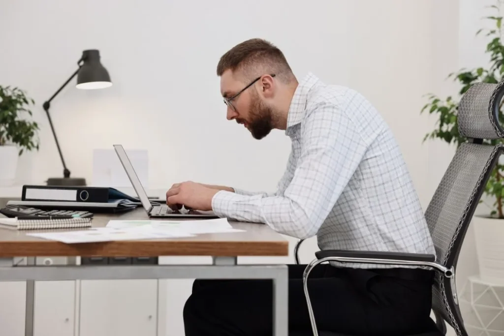 A man sits at a desk working on a laptop while leaning forward with a hunched posture. His head is extended toward the screen and shoulders are rounded, illustrating poor ergonomics and neck strain during typing.
