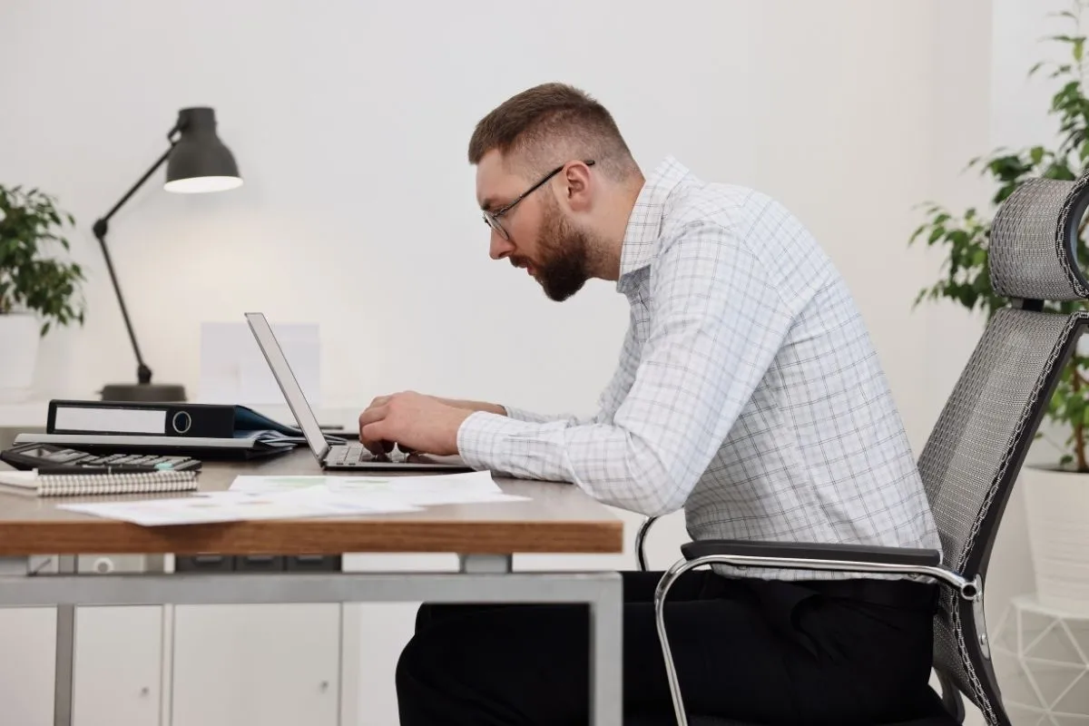 A man sits at a desk working on a laptop while leaning forward with a hunched posture. His head is extended toward the screen and shoulders are rounded, illustrating poor ergonomics and neck strain during typing.