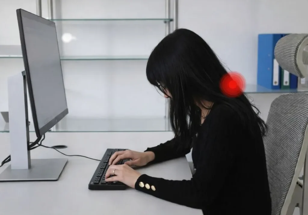 A person sitting at a desk types on a keyboard in front of a computer monitor while leaning forward. Their head is pushed toward the screen and the neck is bent, with a highlighted red area indicating strain in the neck, illustrating “tech neck.”